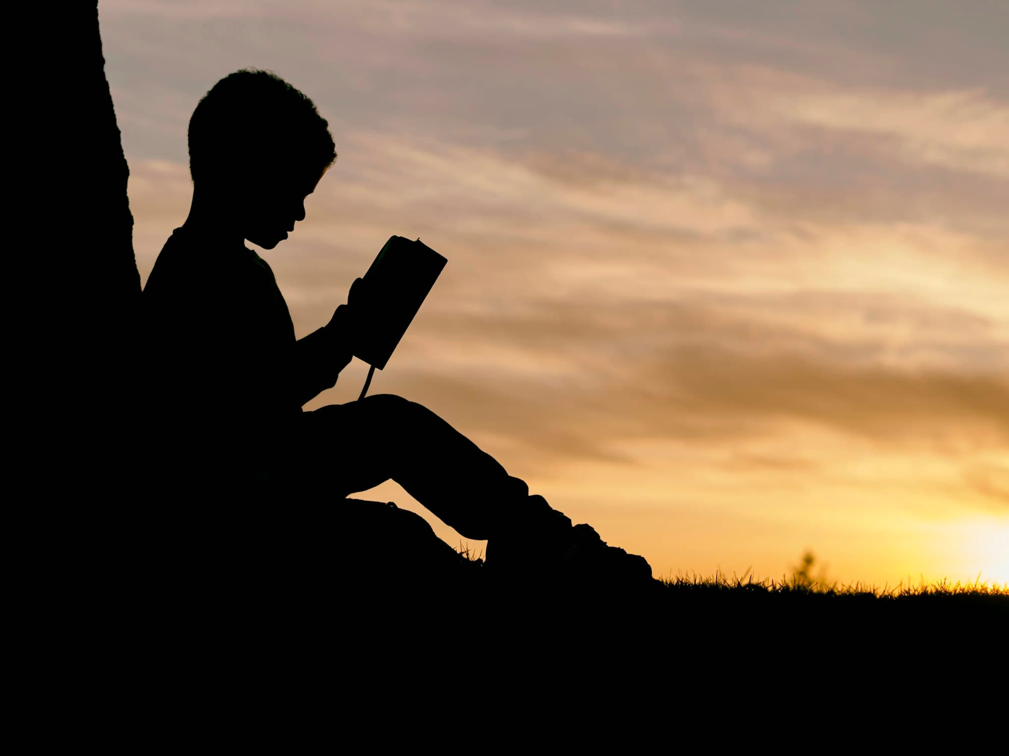 Un enfant lit un livre, en silhouette, dans la lumière dorée du soir.